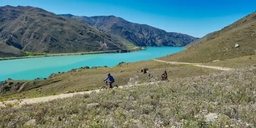 Two cyclists ride along the Lake Dunstan Trail with turquoise waters and rugged mountains in the background