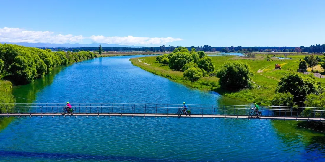 Cyclists crossing a suspension bridge over a calm river on the Nelson to Mapua trail