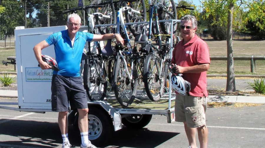 Two cyclists beside a Nelson Cycle Hire trailer loaded with bikes