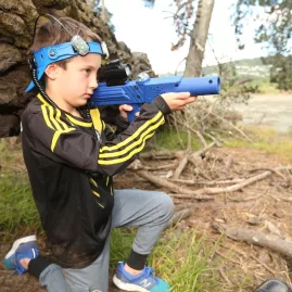Young boy kneeling behind a fallen tree while aiming a blue laser tag gun at Pro Combat Laser Tag in Nelson