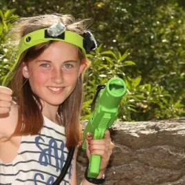 Young girl giving a thumbs up while holding a green laser tag gun during an outdoor game at Pro Combat in Nelson