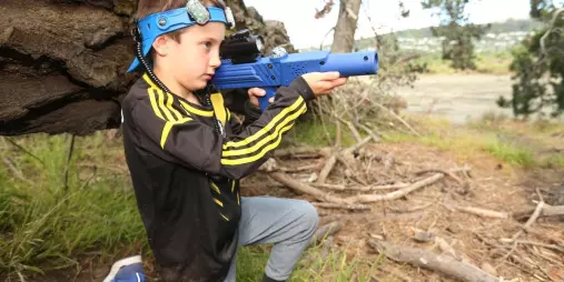 Young boy kneeling behind a fallen tree while aiming a blue laser tag gun at Pro Combat Laser Tag in Nelson