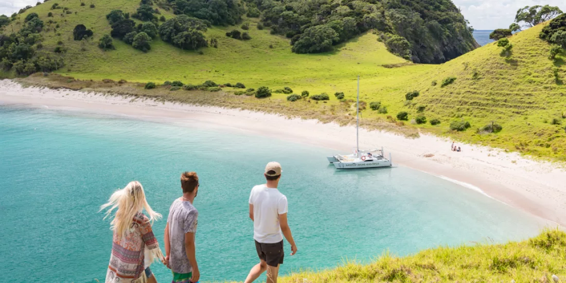 Three travellers walking down a grassy hill toward a catamaran anchored at Waewaetorea Island