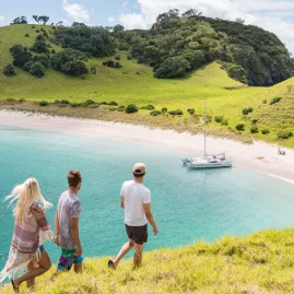 Three travellers walking down a grassy hill toward a catamaran anchored at Waewaetorea Island