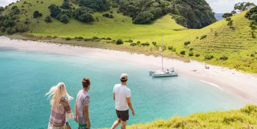 Three travellers walking down a grassy hill toward a catamaran anchored at Waewaetorea Island