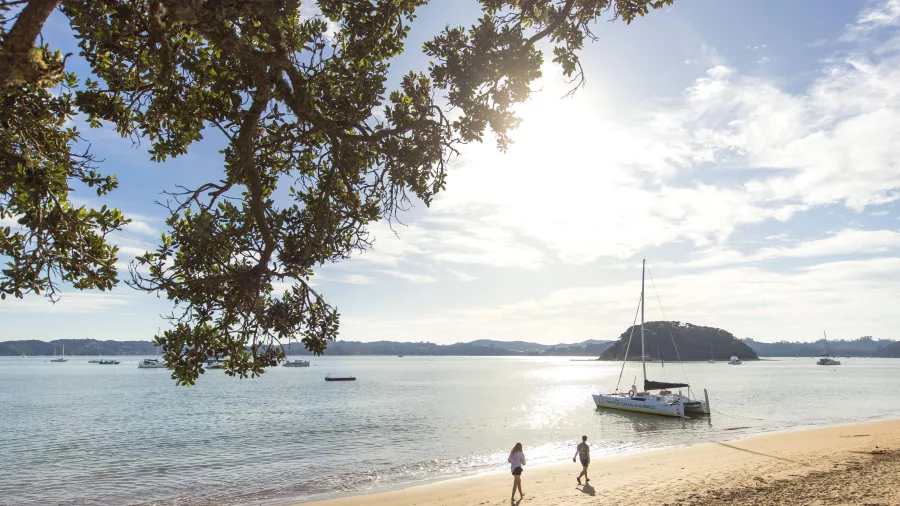 Catamaran anchored near Paihia as two people walk along the beach in the Bay of Islands