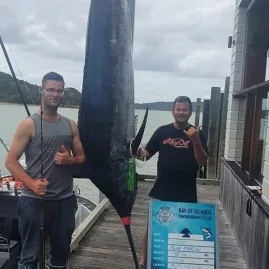 Two men posing beside a large blue marlin at the weigh-in station in the Bay of Islands