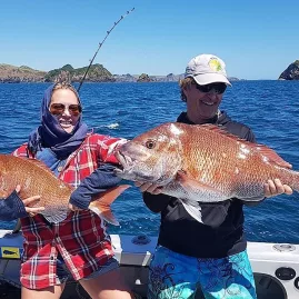 Man and woman holding large snapper on a boat with scenic rock formations in the Bay of Islands