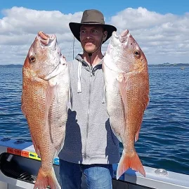 Man in a wide-brimmed hat proudly holding two large snapper fish on a boat in the Bay of Islands