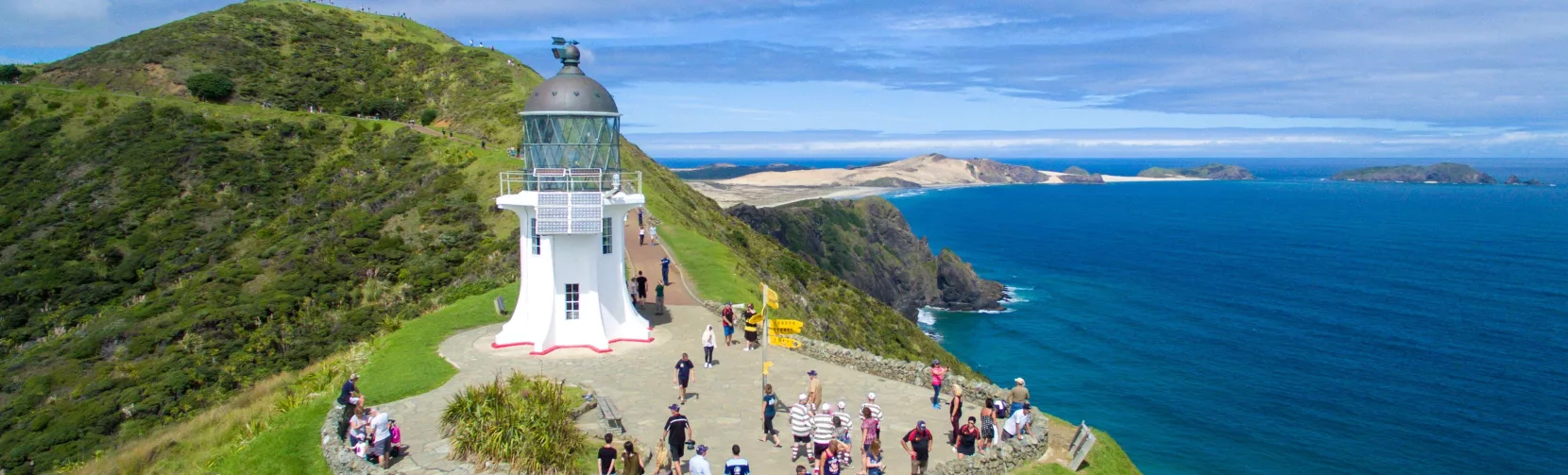 A scenic aerial view of Cape Reinga Lighthouse in Northland, New Zealand, where visitors gather to enjoy panoramic views of the meeting point of the Tasman Sea and the Pacific Ocean.