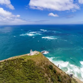 A breathtaking aerial view of Cape Reinga Lighthouse, where the Tasman Sea and Pacific Ocean meet, surrounded by rugged coastal cliffs and lush greenery.