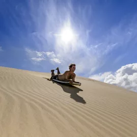 A thrill-seeker sandboarding down the golden Te Paki Sand Dunes in Northland, New Zealand, under a bright blue sky.