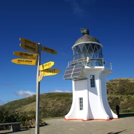 Cape Reinga Lighthouse in New Zealand, standing against a clear blue sky with a yellow directional signpost indicating distances to various world cities.
