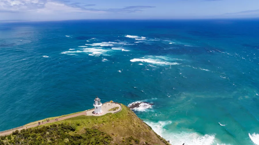 A breathtaking aerial view of Cape Reinga Lighthouse, where the Tasman Sea and Pacific Ocean meet, surrounded by rugged coastal cliffs and lush greenery.