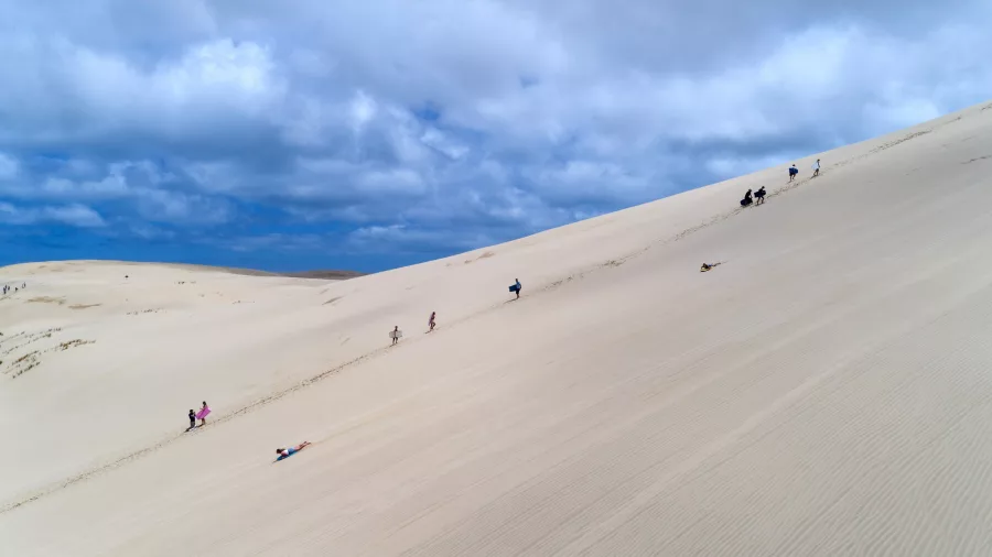 Visitors enjoying sandboarding and hiking on the vast Te Paki Sand Dunes in Northland, New Zealand, under a partly cloudy sky.