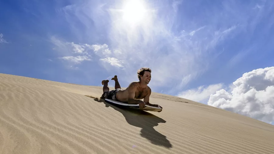 A thrill-seeker sandboarding down the golden Te Paki Sand Dunes in Northland, New Zealand, under a bright blue sky.