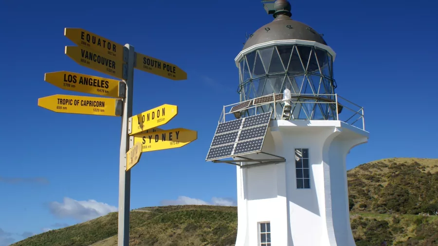 Cape Reinga Lighthouse in New Zealand, standing against a clear blue sky with a yellow directional signpost indicating distances to various world cities.