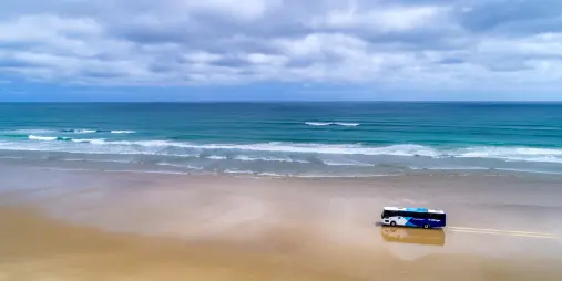 A tour bus driving along the vast, sandy shores of 90 Mile Beach in New Zealand, with ocean waves rolling in under a cloudy sky.