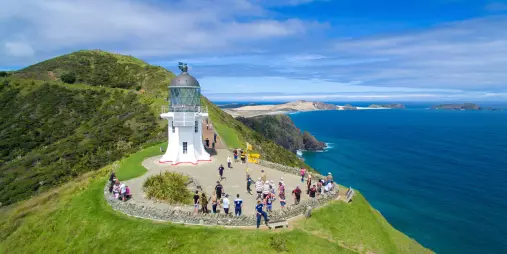 A scenic aerial view of Cape Reinga Lighthouse in Northland, New Zealand, where visitors gather to enjoy panoramic views of the meeting point of the Tasman Sea and the Pacific Ocean.