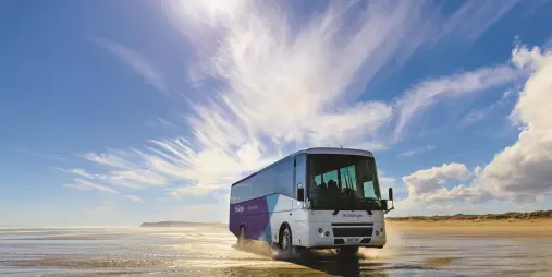 A Fullers GreatSights tour bus driving along the wet sand of Ninety Mile Beach under a bright blue sky in Northland, New Zealand.