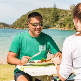 A couple enjoying a delicious meal at a picnic table by the water at Otehei Bay in the Bay of Islands, surrounded by stunning coastal scenery.