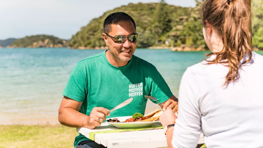 A couple enjoying a delicious meal at a picnic table by the water at Otehei Bay in the Bay of Islands, surrounded by stunning coastal scenery.