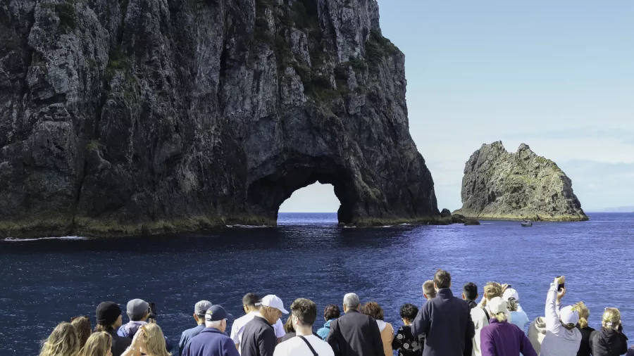 A group of tourists on a boat tour admiring the famous Hole in the Rock formation in the Bay of Islands, surrounded by deep blue waters.