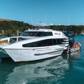 An Explore Group catamaran docked at a pier in the Bay of Islands, New Zealand, with clear blue skies and lush green hills in the background.