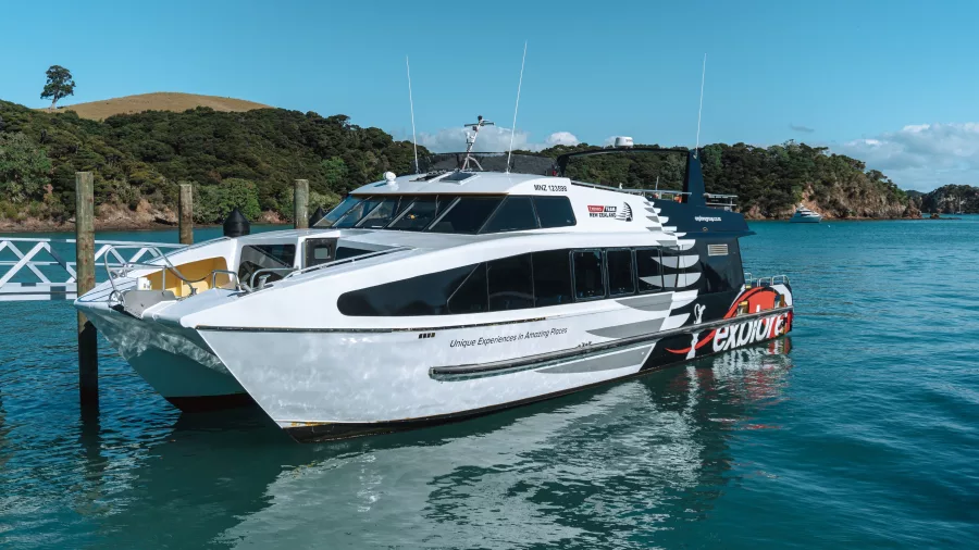 An Explore Group catamaran docked at a pier in the Bay of Islands, New Zealand, with clear blue skies and lush green hills in the background.