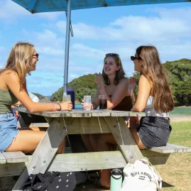 Three young women enjoying a picnic at a wooden table under an umbrella at Otehei Bay, Bay of Islands, New Zealand.