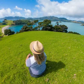 Woman sitting on a green hill overlooking blue waters at Urupukapuka Island, Bay of Islands, New Zealand.