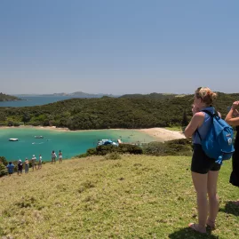 Two hikers with backpacks enjoying a scenic view of Otehei Bay, Bay of Islands, New Zealand, with turquoise waters and a sandy beach below.