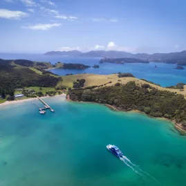Aerial view of Otehei Bay in the Bay of Islands, New Zealand, featuring turquoise waters, a pier, and a boat cruising through.