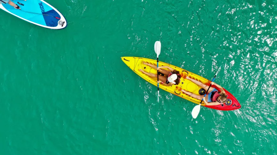 Aerial view of a bright yellow and red kayak with two people paddling, alongside a paddleboarder in turquoise waters of the Bay of Islands, New Zealand.