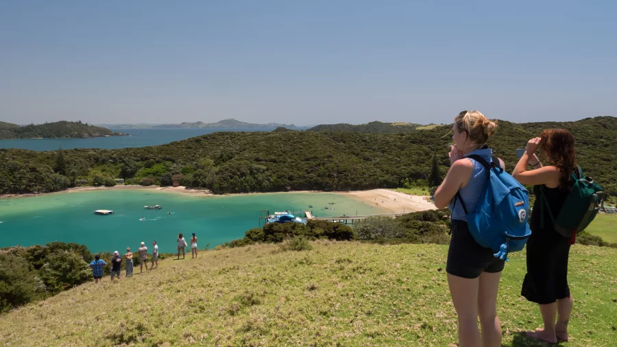 Two hikers with backpacks enjoying a scenic view of Otehei Bay, Bay of Islands, New Zealand, with turquoise waters and a sandy beach below.