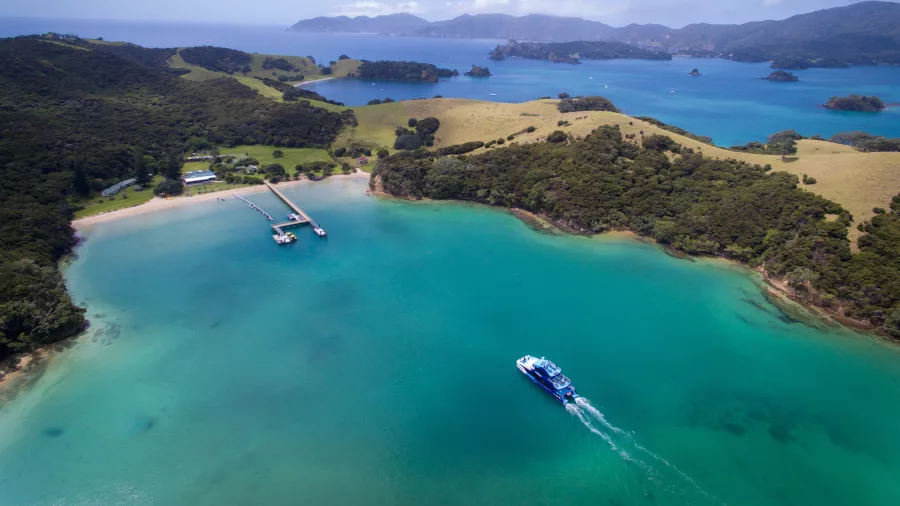 Aerial view of Otehei Bay in the Bay of Islands, New Zealand, featuring turquoise waters, a pier, and a boat cruising through.