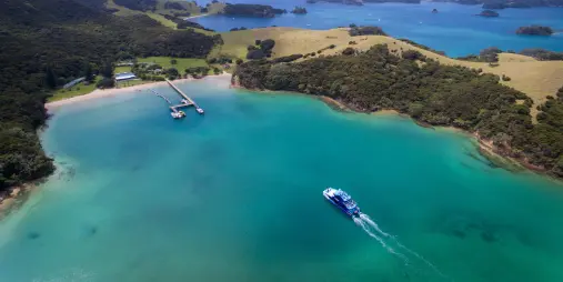 Aerial view of Otehei Bay in the Bay of Islands, New Zealand, featuring turquoise waters, a pier, and a boat cruising through.