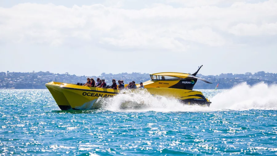 A bright yellow Ocean Adventure speedboat cruising at high speed, creating white waves on the blue waters of New Zealand.
