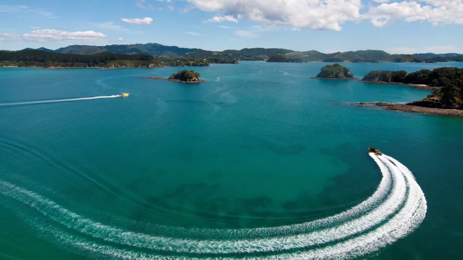 Aerial view of a speedboat making a sharp turn in the turquoise waters of the Bay of Islands, New Zealand, leaving a white wake trail.