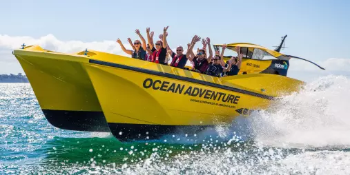 A group of excited passengers enjoying a high-speed Ocean Adventure thrill ride on a bright yellow powerboat in New Zealand.