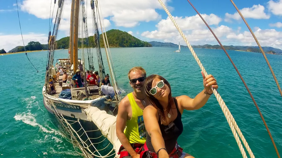 A happy couple taking a selfie on the bowsprit of a sailing ship, surrounded by turquoise waters and tropical islands under a bright blue sky.