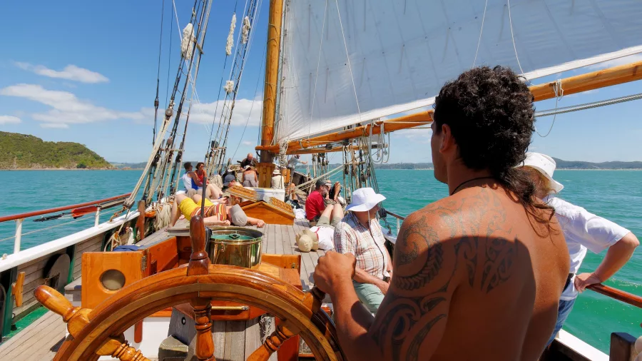 A crew member with a tattooed back steering the R. Tucker Thompson tall ship, while passengers relax on deck under full sails.