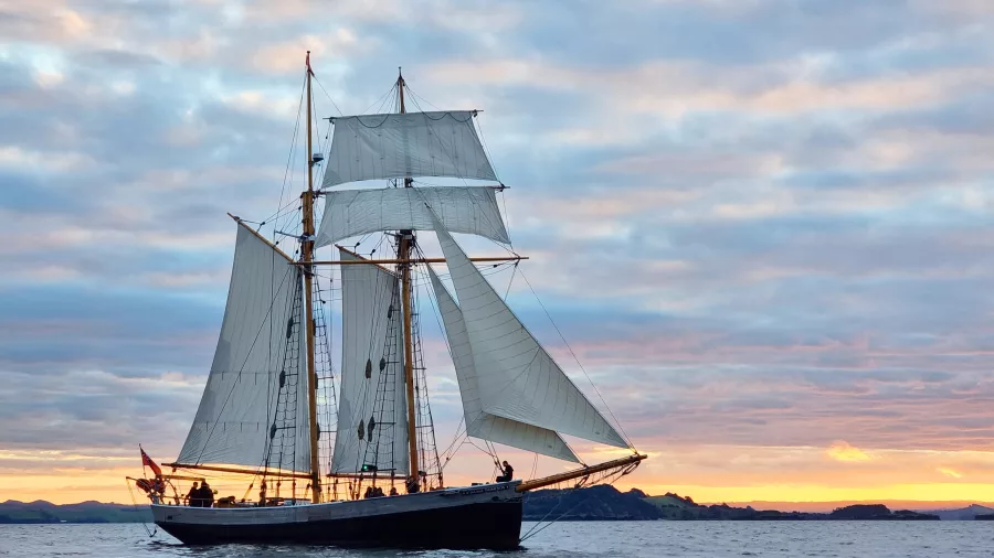 The R. Tucker Thompson tall ship sailing at sunset with full sails against a colorful sky.
