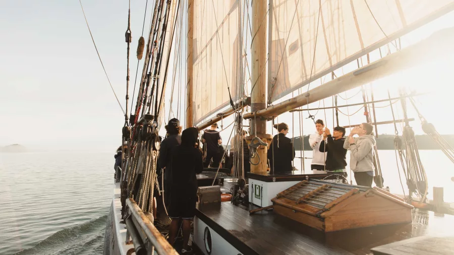 Passengers enjoying a serene sunset sail aboard the R. Tucker Thompson tall ship, with golden light filtering through the sails.