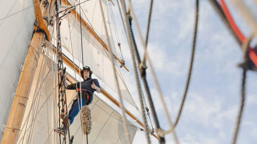 A crew member climbs the rigging of the R. Tucker Thompson tall ship, high above the deck with sails billowing in the wind.