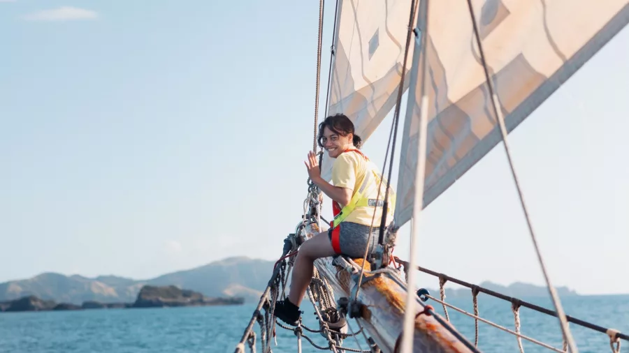 A young passenger sits on the bowsprit of the R. Tucker Thompson, enjoying the view of the Bay of Islands, New Zealand, as the ship sails under clear blue skies.