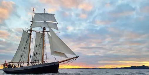 A majestic sailing ship with white sails gliding over the ocean at sunset, with a colorful sky in the background.