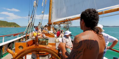 A crew member with a tattooed back steering the R. Tucker Thompson tall ship, while passengers relax on deck under full sails.