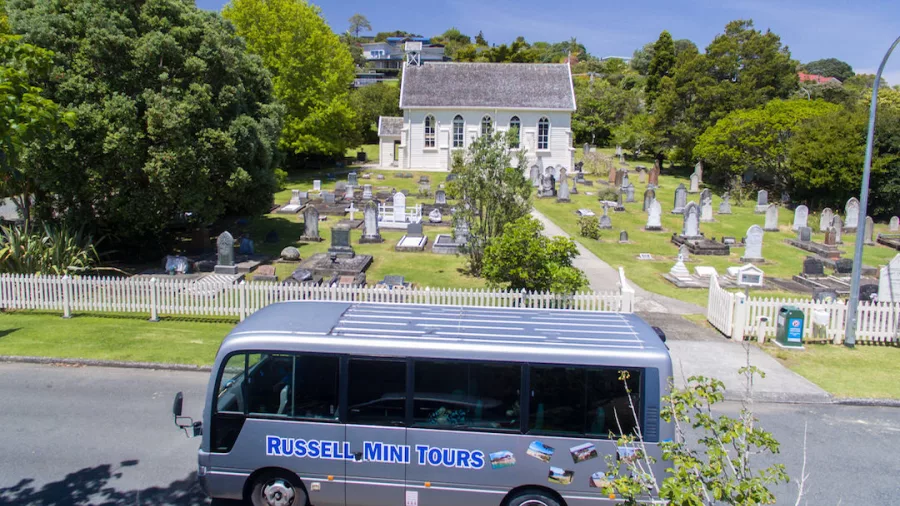 A Russell Mini Tours bus parked outside Christ's Church, a historic church with a graveyard, surrounded by lush greenery on a sunny day.