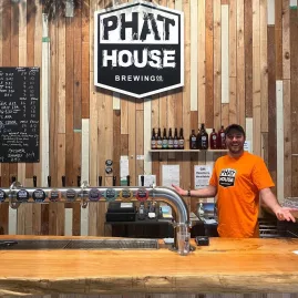 Bartender behind the tap line at Phat House Brewing bar with a wall of craft beer taps and bottles